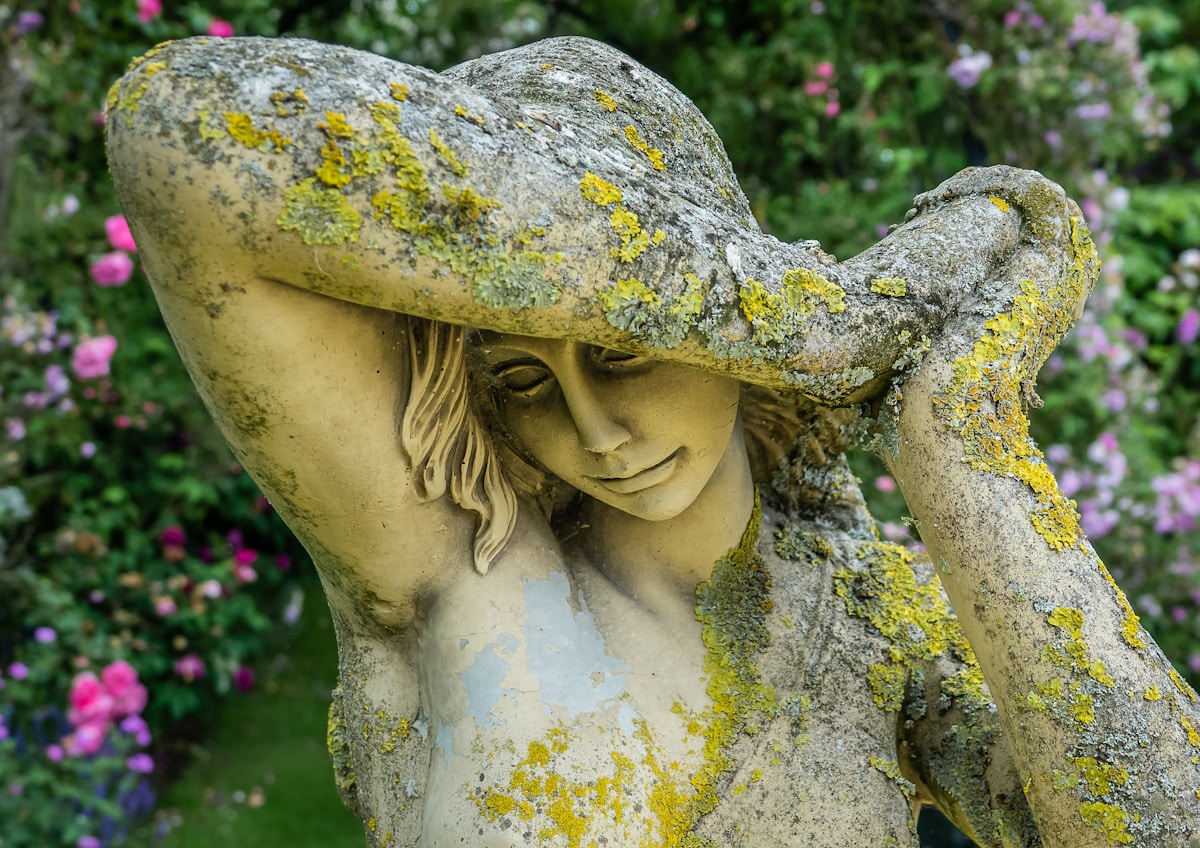 Statue de femme en pierre couverte de mousse et de lichen dans un jardin fleuri.