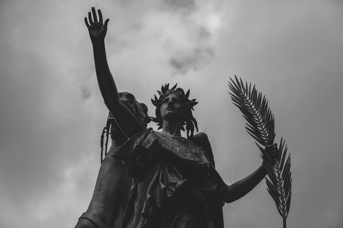 Statue d'une figure féminine avec un bras levé, couronnée de feuilles, contre un ciel nuageux.